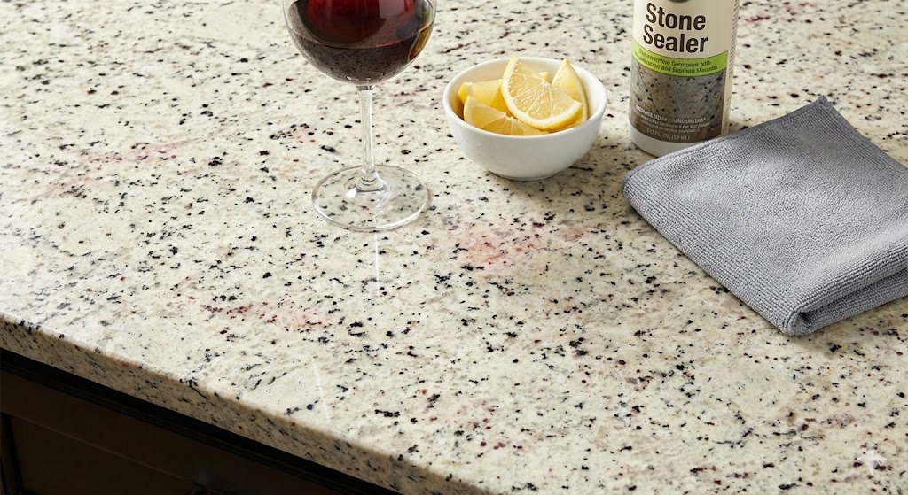 A close-up photograph showing a Colonial White granite countertop with a bottle of stone sealer, a microfiber cloth, a glass of red wine, and a bowl of lemons, illustrating proper maintenance and stain prevention.