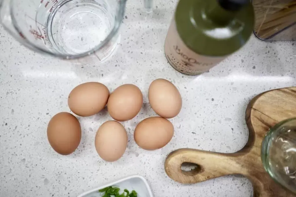 “Six brown eggs on a white quartz countertop with olive oil, measuring cup, and cutting board. Kitchen prep scene by Granite Guy Inc.”