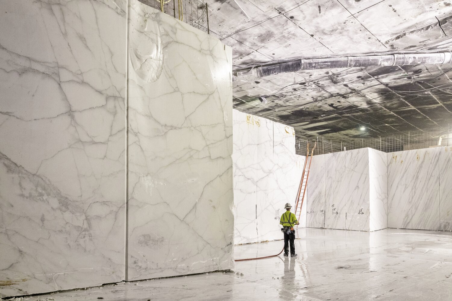 A massive underground marble quarry showing large blocks of Calacatta Lincoln marble with a worker in high-visibility gear for scale. The scene highlights the impressive extraction process of high-quality white marble used for premium countertops.